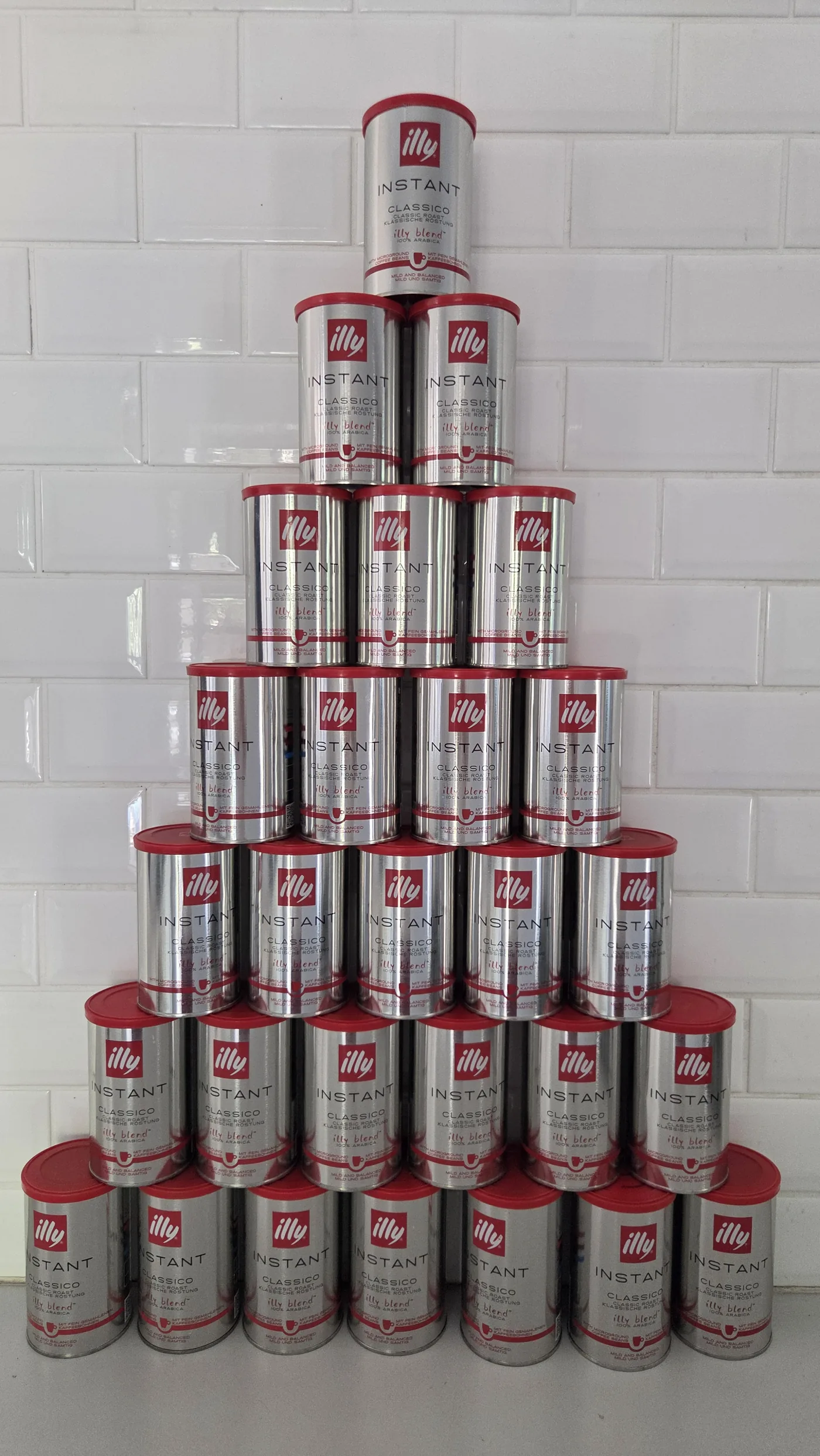 Twenty-eight empty tins of instant coffee stacked in a pyramid against white tiles